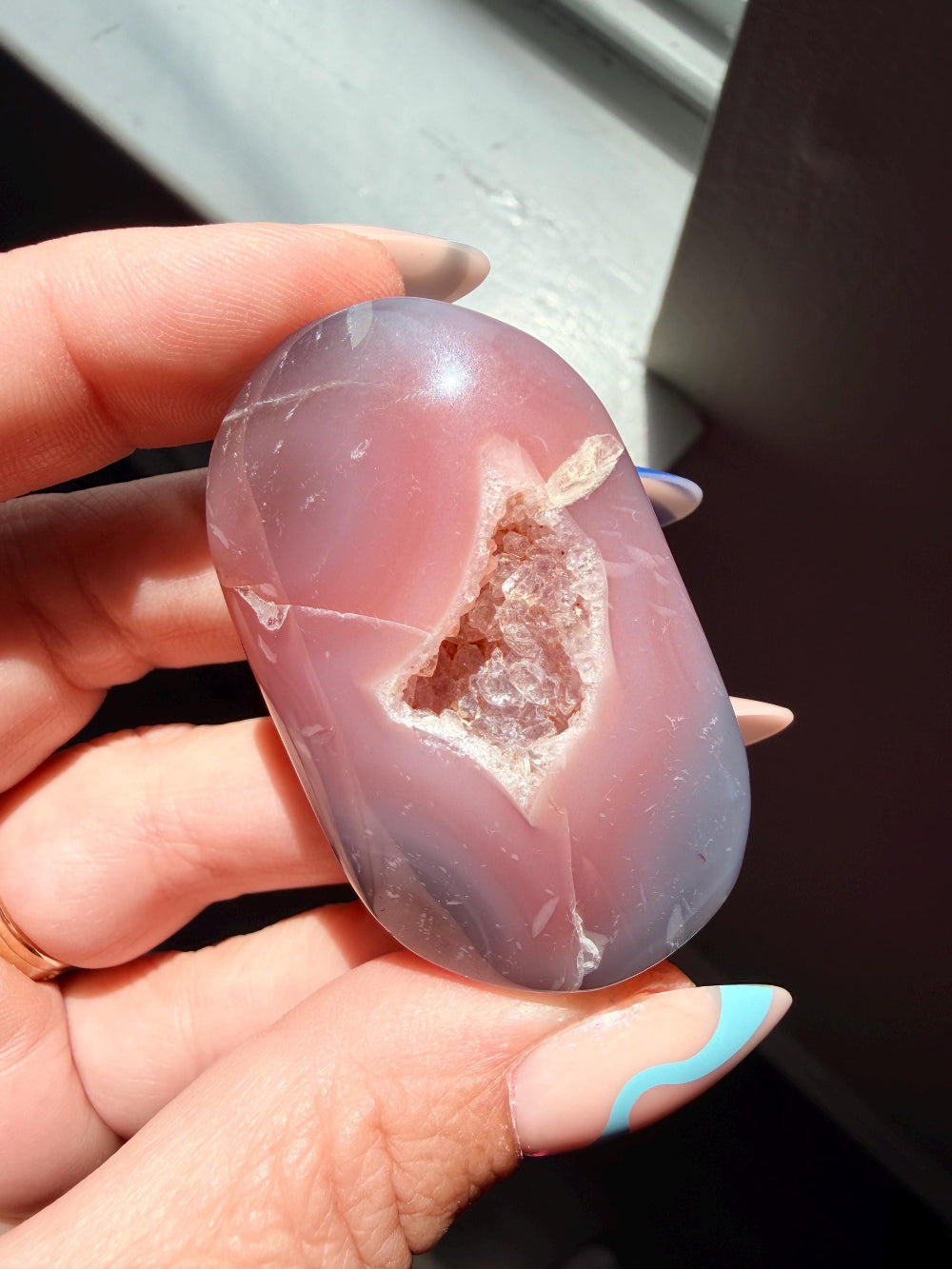 A hand holding a polished oval agate stone with a natural geode cavity filled with brown crystal formations. The stone has a dusty rose and lavender-purple coloration with white veining.